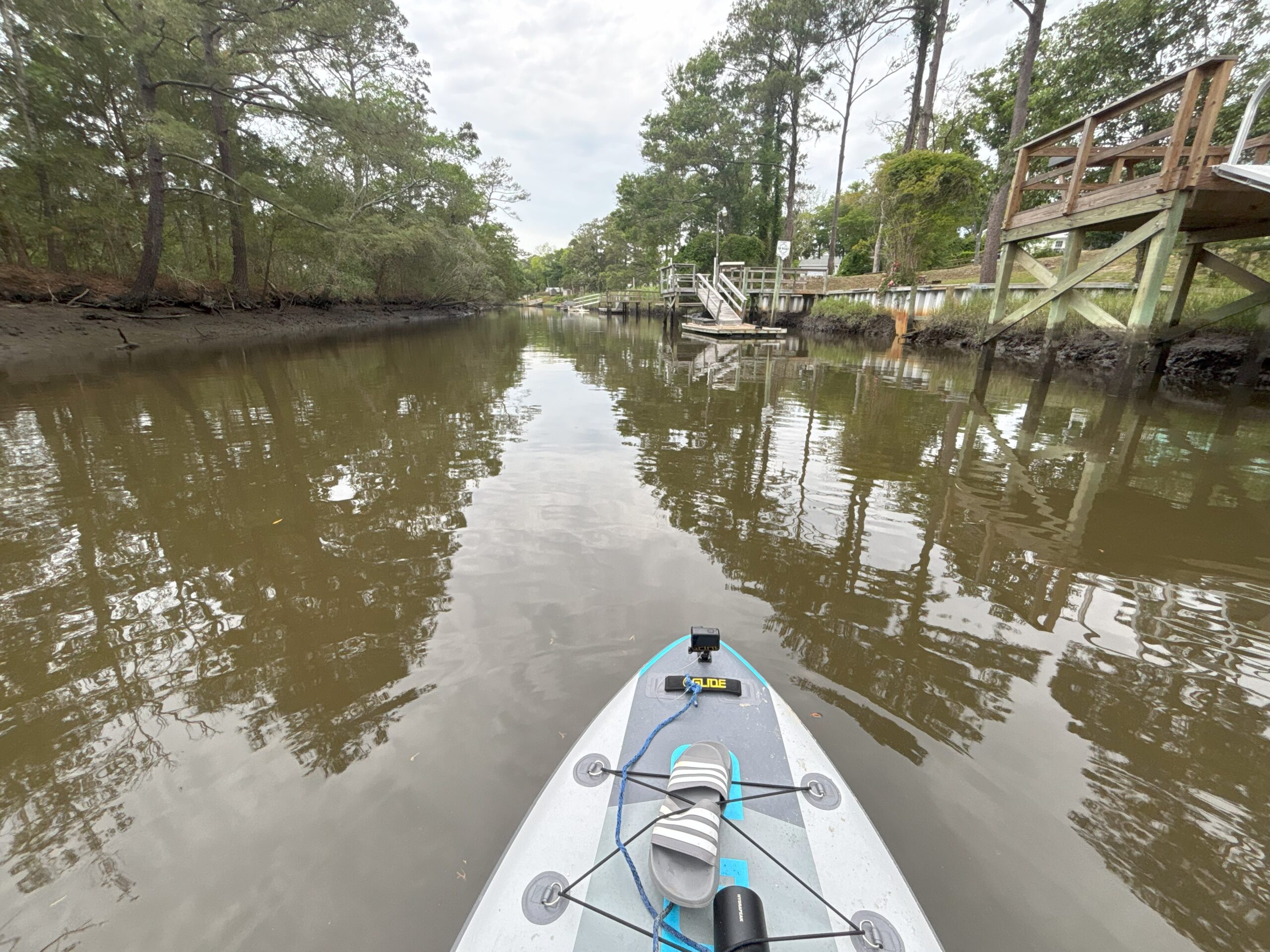 person's POV being on a paddleboard on the oak island canal with brown water, trees and docks nearby.
