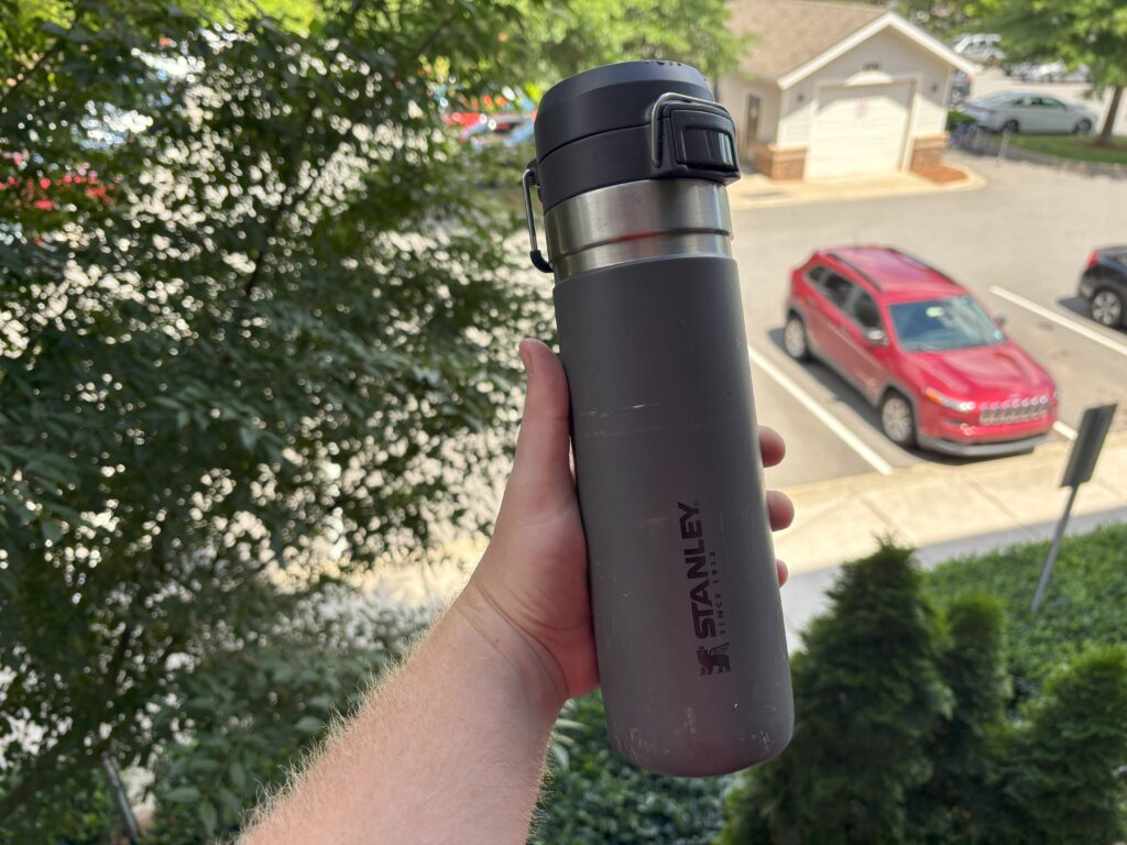 My Stanley Quick Flip GO that has taken a beating. Behind it is a parking lot and tree. It still is in functioning shape as a thermos for camping.