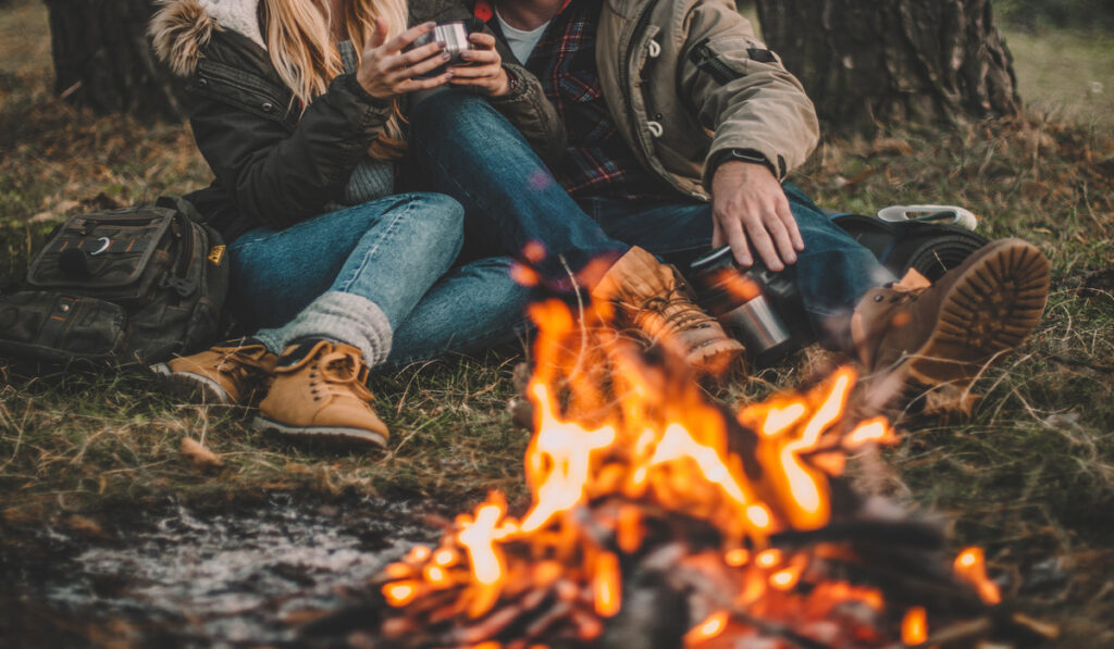 A man and woman couple in warm clothing sitting near a campfire outside while drinking from aluminum mugs.