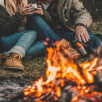 A man and woman couple in warm clothing sitting near a campfire outside while drinking from aluminum mugs.
