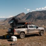 A truck with a pile of equipmen tin the bed is parked on a patch of dirt. In the distance are barren mountains.