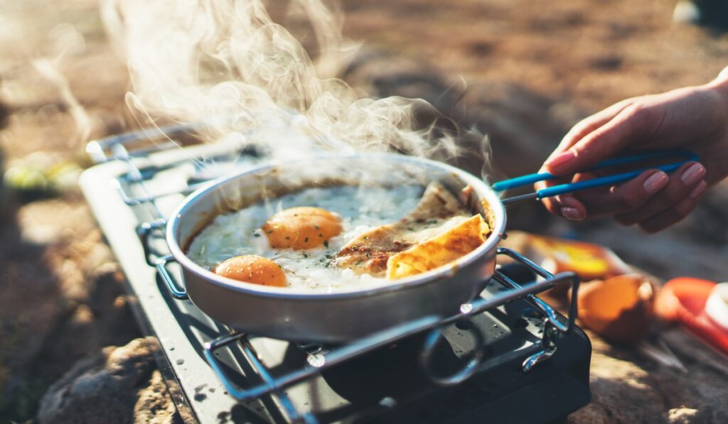 A close-up of a frying pan on a portable stove outdoors, cooking eggs, meat, and bread, with steam rising visibly.