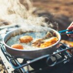 A close-up of a frying pan on a portable stove outdoors, cooking eggs, meat, and bread, with steam rising visibly.