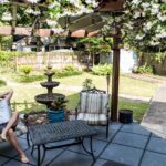 A man lounging with his arms behind his head on an outdoor armchair on his patio. A pergola covered in white blooms is above him.