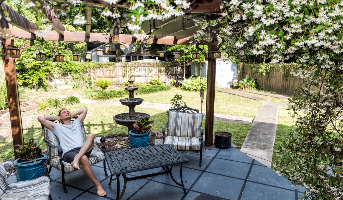 A man lounging with his arms behind his head on an outdoor armchair on his patio. A pergola covered in white blooms is above him.